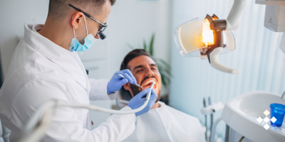 A dentist wearing a mask and loupes examines a patient's teeth with a dental tool.